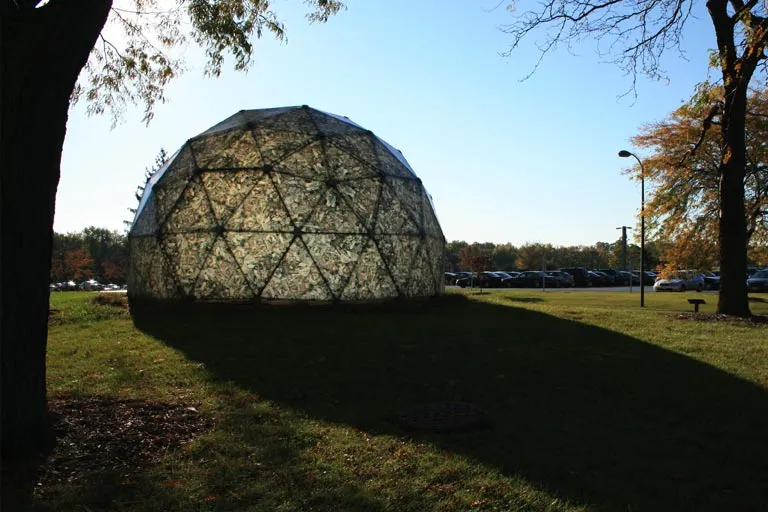Large translucent geodesic dome on green grass, backlit by sun, with autumn trees.