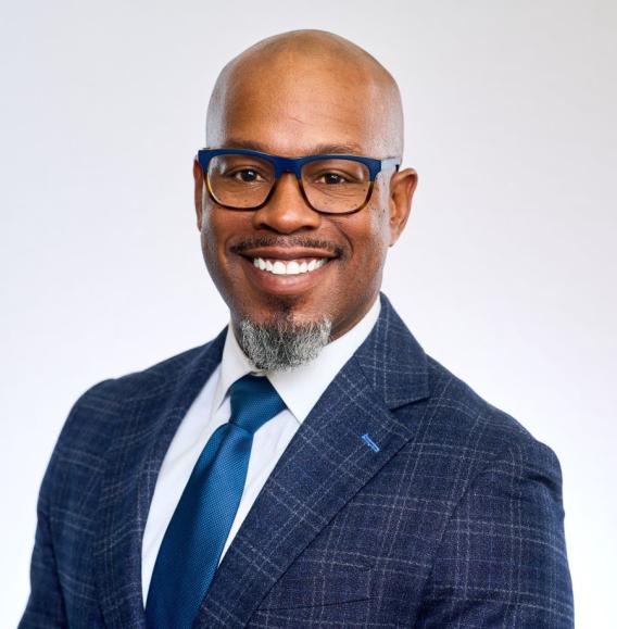 Smiling man in blue plaid suit, blue tie, blue glasses, and gray goatee.